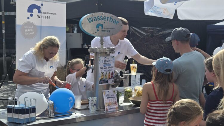 Mitarbeitende von RheinHunsrück Wasser schenken an der Wasserbar Trinkwasser mit frischen Zutaten aus, Kinder und Besucher stehen am Tresen.